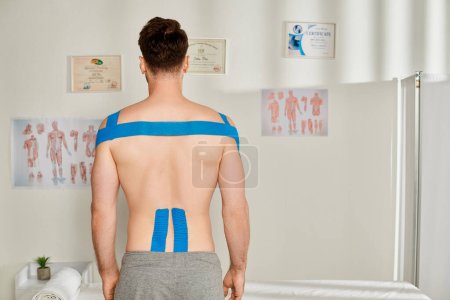 Rückenansicht des männlichen Patienten in grauer Jogginghose mit kinesiologischen Klebebändern am Körper, Gesundheitswesen
