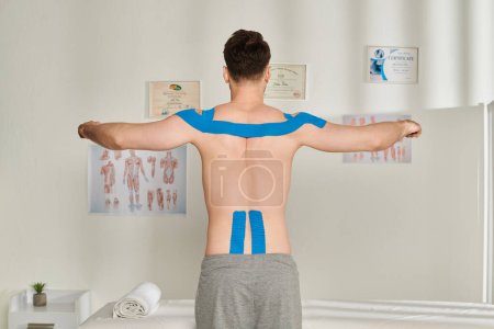Téléchargez les photos : Vue arrière du patient masculin posant avec les bras de côté avec des bandes kinésiologiques sur son corps, les soins de santé - en image libre de droit