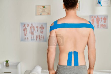 Téléchargez les photos : Vue arrière du patient masculin pendant le rendez-vous avec des bandes kinésiologiques sur son dos et ses épaules - en image libre de droit