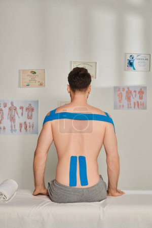 Téléchargez les photos : Vue arrière de l'homme assis lors d'un rendez-vous avec des bandes kinésiologiques sur le dos, soins de santé - en image libre de droit