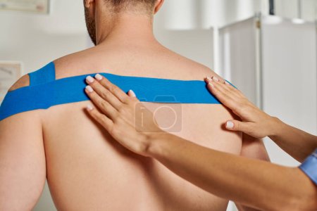 Ausgeschnittene Ansicht der Hände einer jungen Ärztin, die ihrem männlichen Patienten kinesiologisches Klebeband auf den Rücken legt