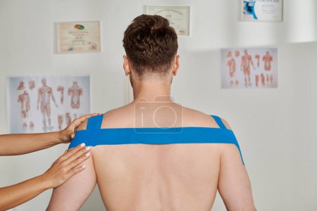 back view of man with hand of female woman putting kinesiological tapes on his shoulders and back