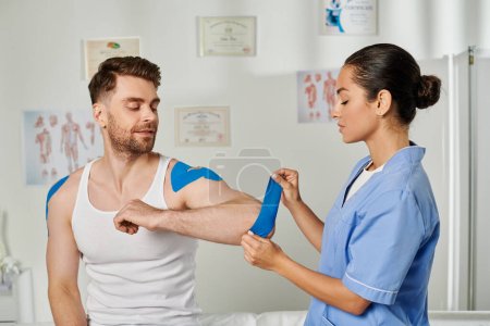 Photo for Jolly handsome patient sitting and watching his doctor putting kinesiological tape on his elbow - Royalty Free Image