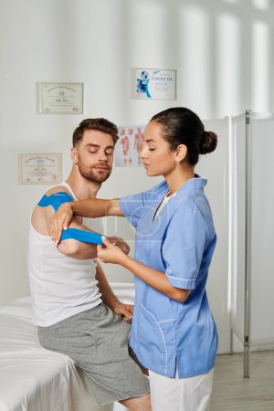 Foto de Atractiva joven médico en traje médico poniendo cinta de kinesio en su paciente guapo, cuidado de la salud - Imagen libre de derechos