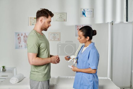 Foto de Buen paciente barbudo mirando atentamente el modelo de columna vertebral en manos de su atractivo médico - Imagen libre de derechos