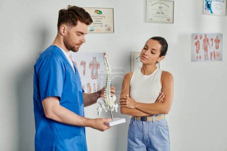 attraktive Patientin beim Anblick eines gutaussehenden bärtigen Arztes mit Wirbelsäulenmodell in seinen Händen