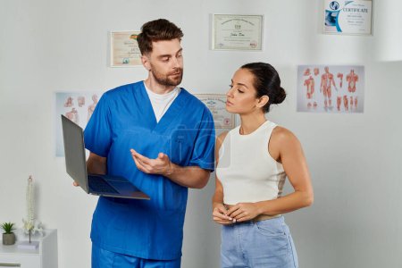 Téléchargez les photos : Beau médecin barbu regardant son patient qui regarde l'ordinateur portable pendant le rendez-vous, les soins de santé - en image libre de droit