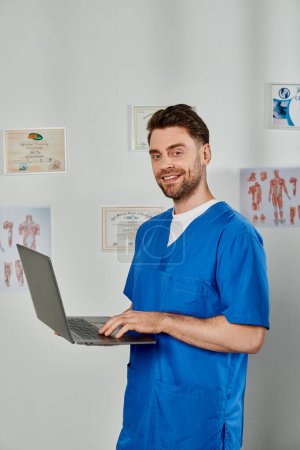 fröhlich gut aussehender Arzt posiert mit Laptop in den Händen und schaut in die Kamera, Gesundheitswesen