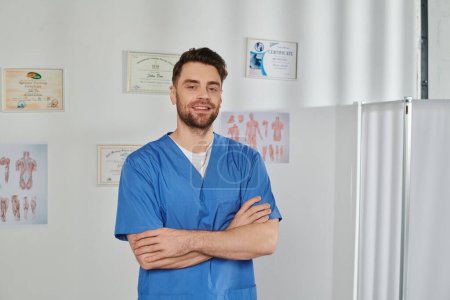 Foto de Alegre médico guapo posando con los brazos cruzados en el pecho y sonriendo a la cámara, atención médica - Imagen libre de derechos