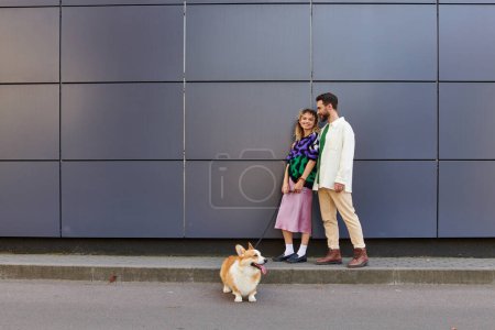 happy and stylish couple walking with corgi dog near modern grey building, animal companions