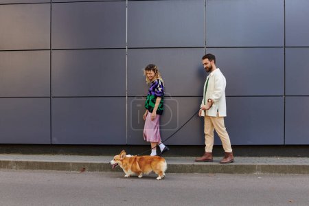 happy and stylish couple walking with corgi dog near modern grey building, animal companions