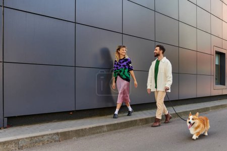 happy couple holding hands and walking with corgi dog near modern grey building, animal companions