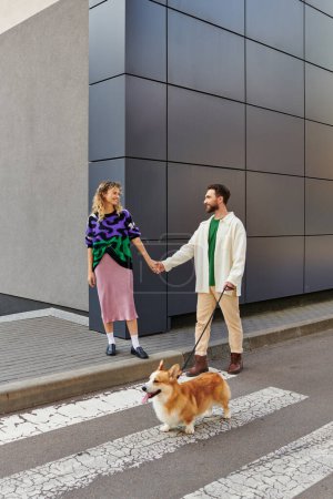 happy couple holding hands and walking with corgi dog near modern grey building, animal companions