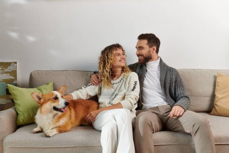 Foto de Feliz pareja en casual invierno trajes sentado en sofá y abrazo corgi perro en moderno apartamento - Imagen libre de derechos