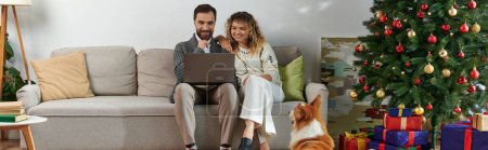 Téléchargez les photos : Bannière, homme utilisant un ordinateur portable et assis sur le canapé avec femme bouclée près de chien de corgi mignon et arbre de Noël - en image libre de droit