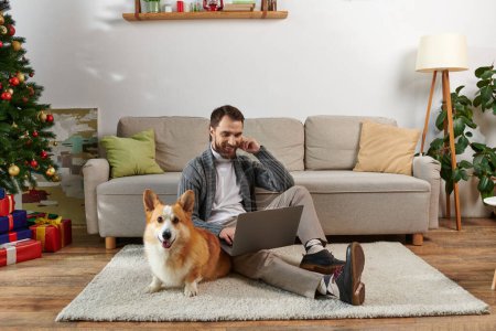 happy man using laptop and sitting on carpet near corgi dog and decorated Christmas tree at home