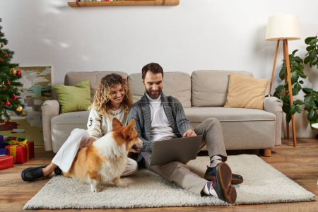 bearded man using laptop and sitting on carpet with curly wife near corgi dog and Christmas tree