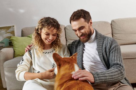 fröhliches Paar lächelt und spielt mit niedlichem Corgi-Hund in moderner Wohnung, glückliche Momente