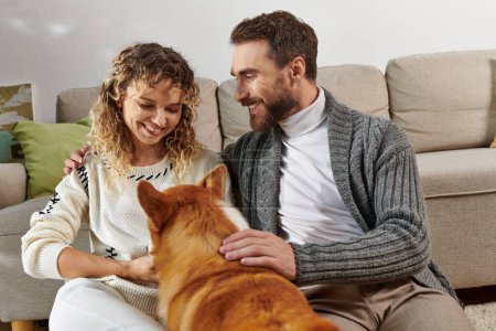 glückliches Paar lächelt und spielt mit süßem Corgi-Hund in moderner Wohnung, glückliche Momente