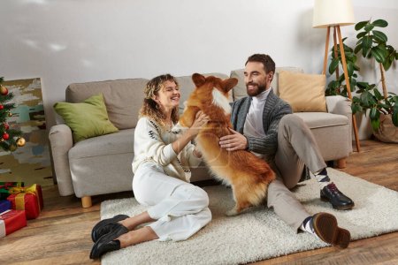 fröhlicher Mann und Frau lächelnd und spielend mit niedlichem Corgi-Hund in moderner Wohnung, glückliche Momente
