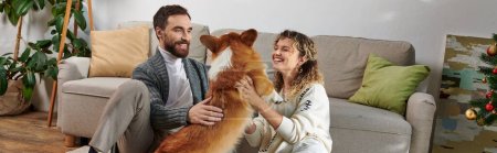 glückliches Paar lächelt und spielt mit niedlichen Corgi-Hund in moderner Wohnung, gemütliche Momente Banner