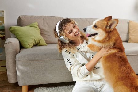 lockige Frau mit drahtlosen Kopfhörern spielt mit niedlichem Corgi-Hund in moderner Wohnung, glückliche Momente