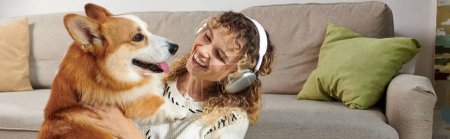 femme bouclée dans des écouteurs sans fil jouer avec chien corgi mignon dans un appartement moderne, moments heureux