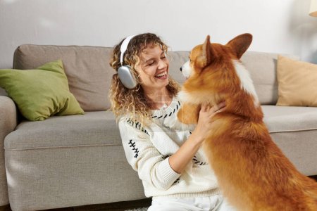 Téléchargez les photos : Femme bouclée dans des écouteurs sans fil jouer avec chien corgi mignon dans un appartement moderne, moments heureux - en image libre de droit