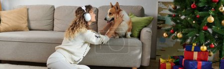 curly woman in wireless headphones playing with cute corgi dog near decorated Christmas tree, banner