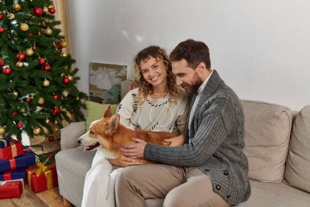 freudiges Paar in Winterkleidung lächelt und spielt mit Corgi-Hund in der Nähe des geschmückten Weihnachtsbaums