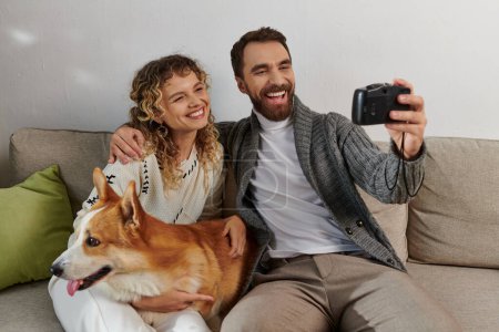 pareja sonriendo y tomando fotos en cámara con perro corgi en apartamento moderno, momentos felices