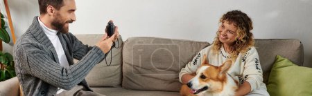 Foto de Feliz hombre sonriendo y tomando la foto de la esposa con el perro corgi en la cámara en el apartamento moderno, pancarta - Imagen libre de derechos
