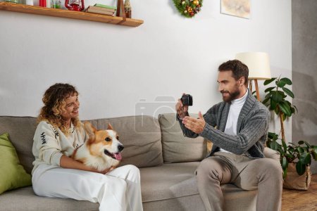 Foto de Hombre sonriendo y tomando la foto de la esposa con el perro corgi en la cámara en el apartamento moderno, momentos felices - Imagen libre de derechos