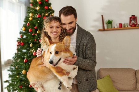 happy couple in winter clothing holding corgi dog and standing near decorated Christmas tree