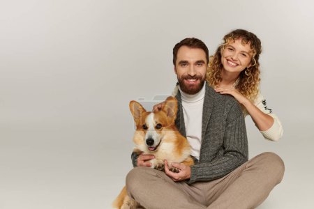 Photo for Family portrait, happy man and woman in winter attire posing with cute corgi on grey background - Royalty Free Image