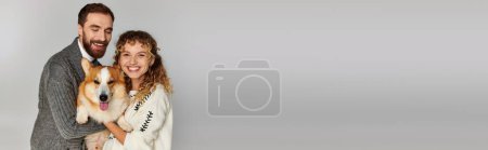 Photo for Family portrait, positive man and woman in winter attire posing with cute corgi on grey background - Royalty Free Image