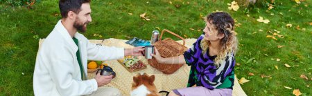 happy curly woman and cheerful man having picnic near cute corgi dog on green lawn in park, banner