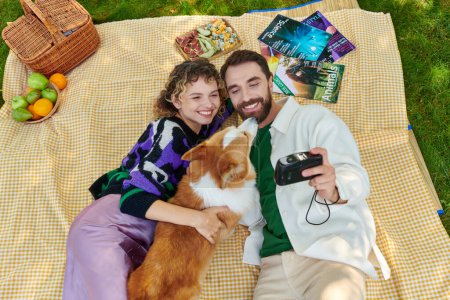 top view of happy couple taking photo with cute corgi dog on camera while having picnic in park