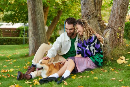 feliz hombre abrazando a la mujer rizada en lindo traje mientras acaricia perro corgi en el parque, sentado cerca del árbol