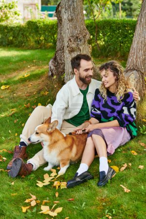 happy man hugging curly woman in cute outfit while cuddling corgi dog in park, sitting near tree