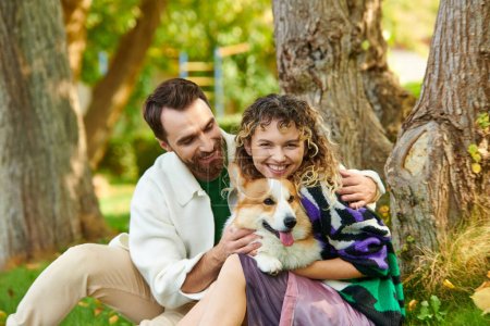 glücklicher Mann umarmt Frau im niedlichen Outfit, während er Corgi-Hund im Park knuddelt, neben Baum sitzend
