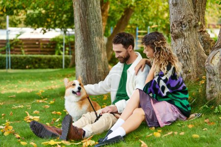 glücklicher Mann und lockige Frau im niedlichen Outfit, die Corgi-Hund anschauen und neben Baum im herbstlichen Park sitzen