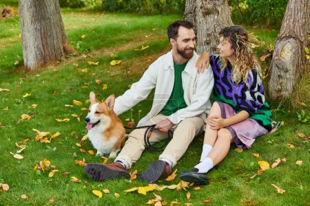 glücklicher Mann schaut lockige Frau im niedlichen Outfit an, die mit Corgi-Hund am Baum im herbstlichen Park sitzt