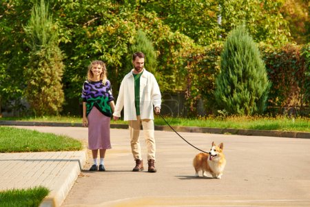 happy couple in stylish attire holding hands and walking with cute corgi dog around green trees