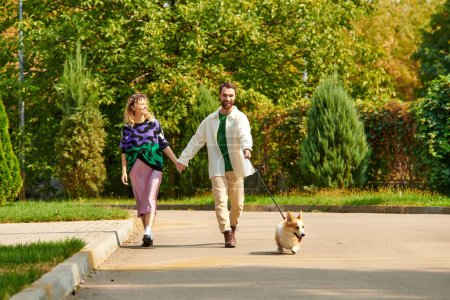 happy couple in stylish attire holding hands and walking with cute corgi dog around green trees