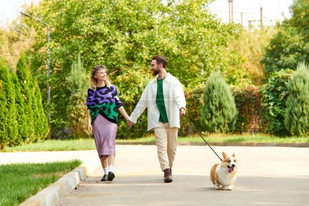 glückliches Paar in stylischer Kleidung, Händchen haltend und mit niedlichem Corgi-Hund um grüne Bäume spazierend