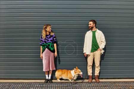 happy and stylish couple walking with corgi dog near grey garage door, animal companions