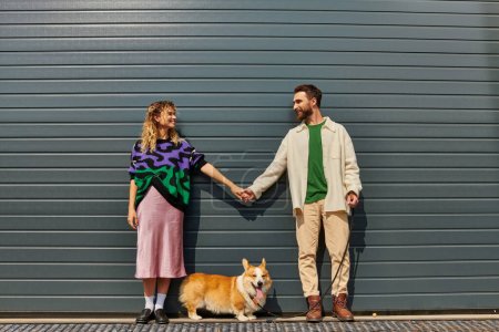 glückliches und stylisches Paar hält Händchen und steht mit Corgi-Hund neben grauem Garagentor