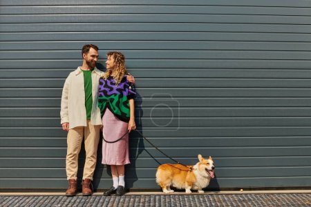 fröhliches und stilvolles Paar, das mit Corgi-Hund in der Nähe von grauen Garagentoren spazieren geht, tierische Begleiter