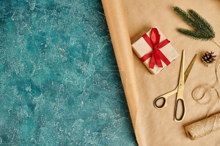 boîte cadeau décorée sur papier artisanal et ciseaux près de ficelle, cône de pin et branche de sapin sur fond bleu